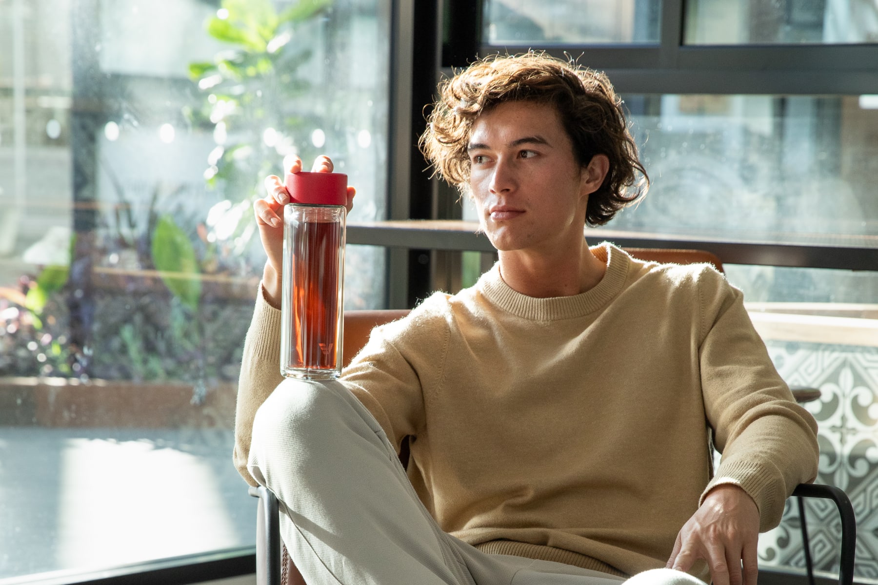 A male holding an Ever Vessel Glass Multi tea flask bottle with red colored lid and black infused tea inside on his knee.