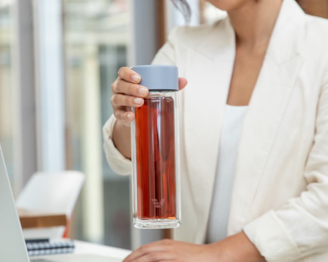 Female lifting an Ever Vessel Glass Multi double wall glass tea flask bottle with lilac colored lid and black infused tea inside at her desk.