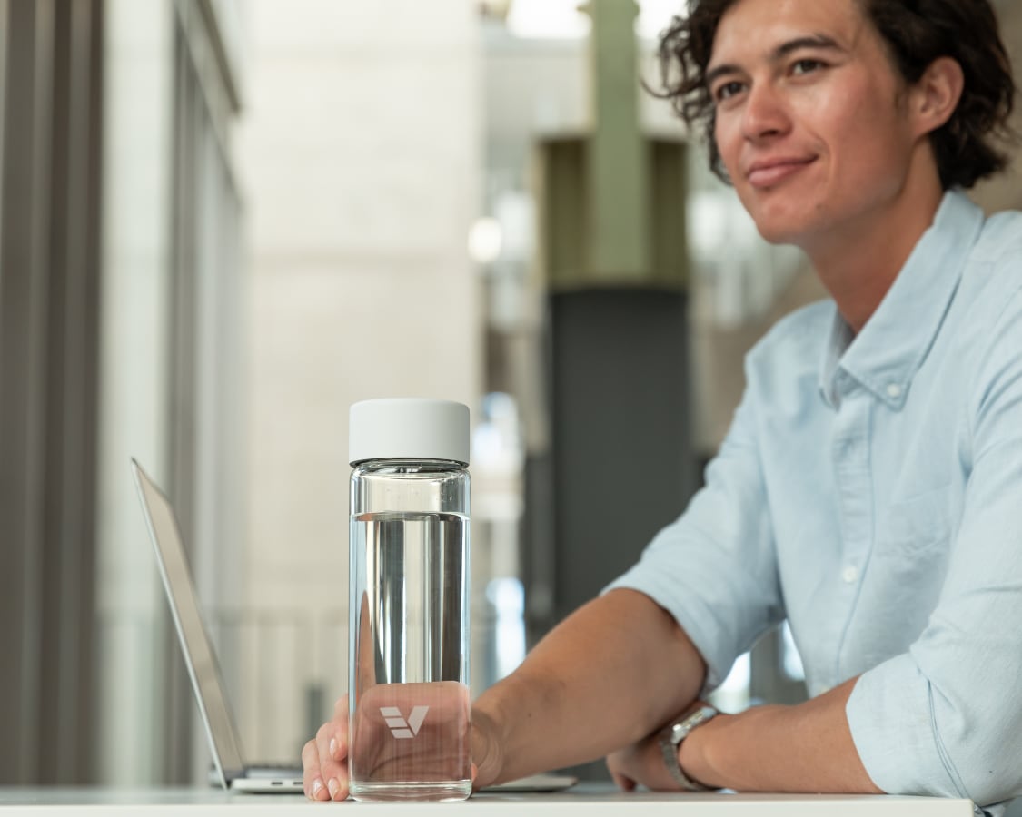A male picking up his Ever Vessel Glass water bottle with a white lid off his desk.