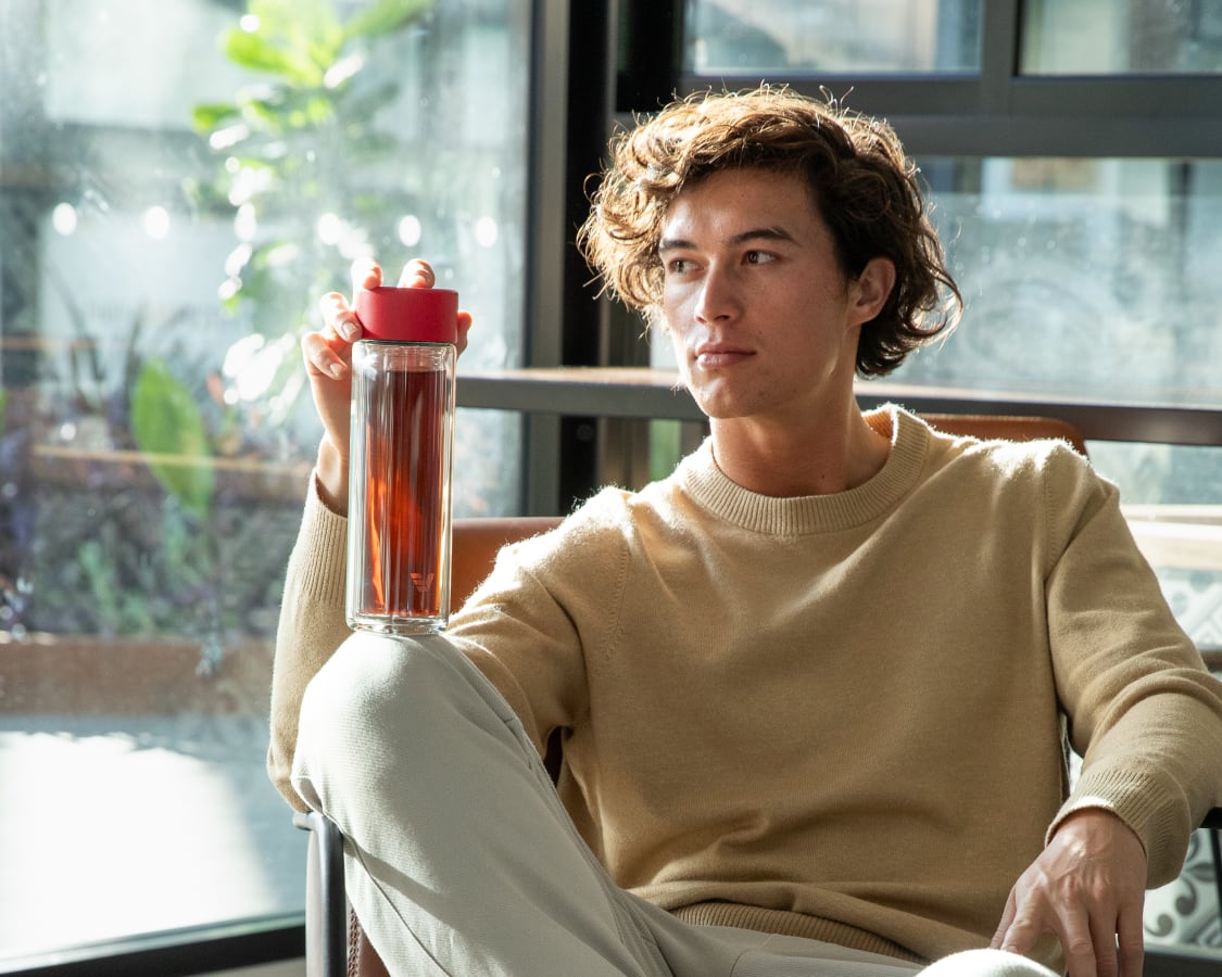 A male holding an Ever Vessel Glass Multi tea flask bottle with red colored lid and black infused tea inside on his knee.