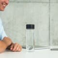 An Ever Vessel Glass water bottle with black lid on an open plan work desk next to a person talking to a work colleague.