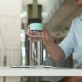 A male picking up his Ever Vessel Glass Multi tea infuser water bottle with a mint green lid off his desk.