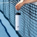 A female holding her Ever Vessel Maxi White Sand stainless steel water bottle by the integrated handle over the tennis court net.