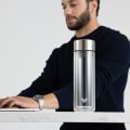 An Ever Vessel Glass Multi  double walled glass tea infuser water bottle with stainless grey lid sitting on a desk next to a male working.