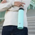 A male holding his Super Maxi Mint stainless steel water bottle by the integrated carry handle on the stairs outside his office.