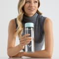 A female holding her Ever Vessel Glass water bottle with mint green lid above a stone bench at work.