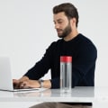 An Ever Vessel Glass Multi double wall glass tea infuser water bottle with red lid on stone desk next to a male working on his laptop.