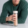 A male holding an Ever Vessel Glass Multi double walled tea flask bottle with Desert beige colored lid on a kitchen bench.