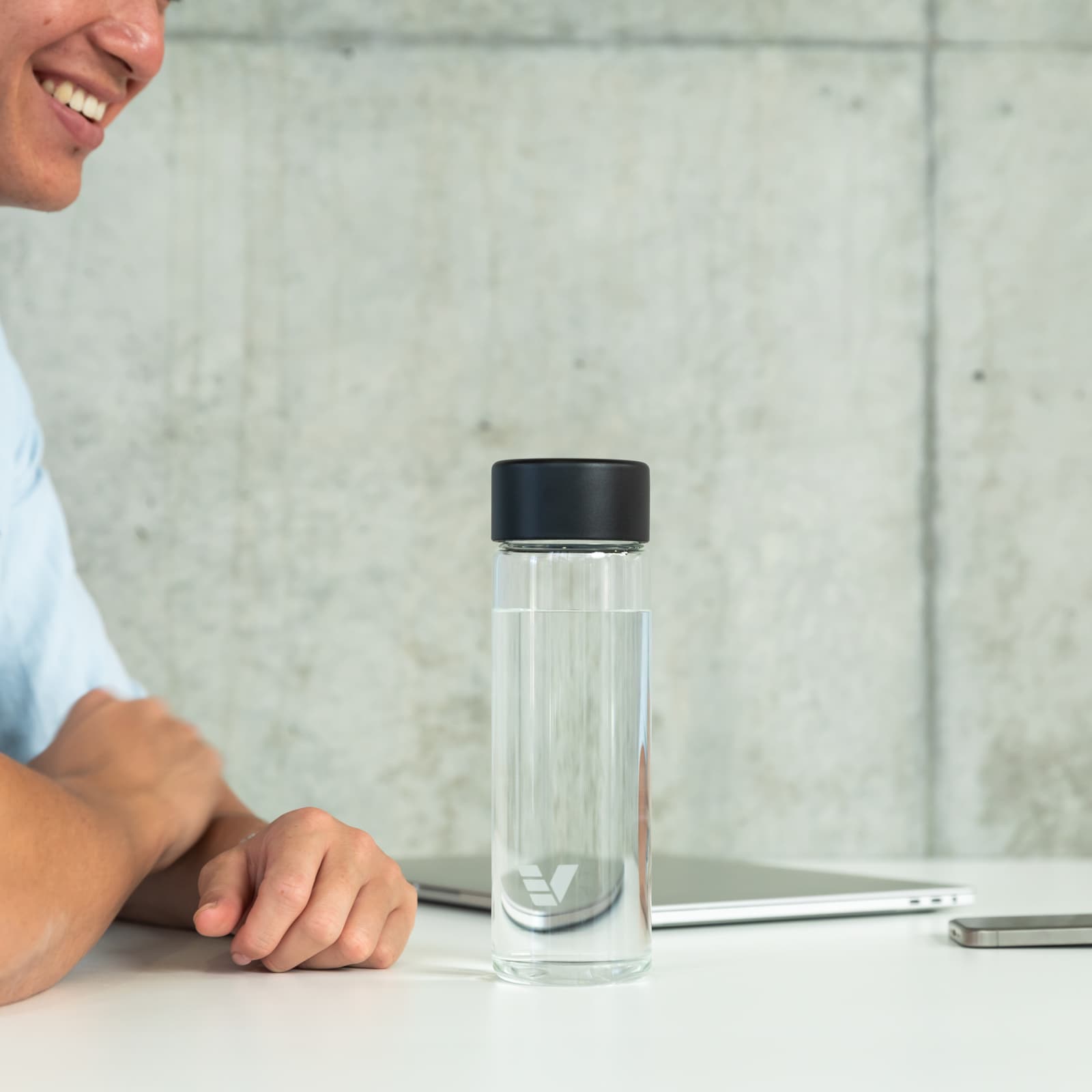 An Ever Vessel Glass water bottle with black lid on an open plan work desk next to a person talking to a work colleague.