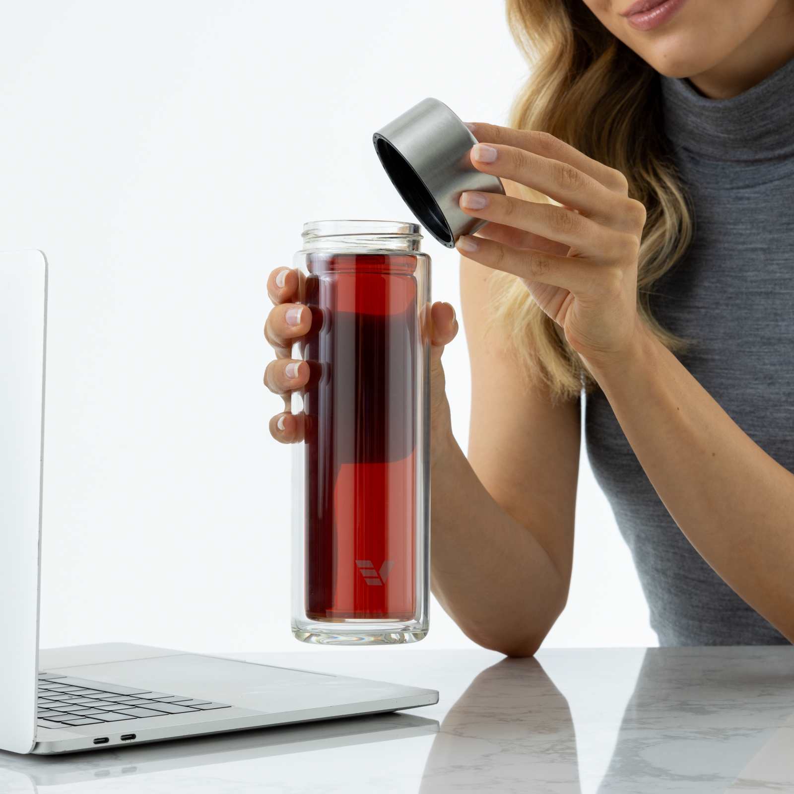A female taking off the stainless grey lid from her Ever Vessel Glass Multi double walled glass tea infuser water bottle at her desk.