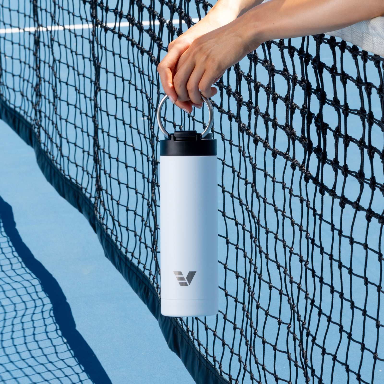 A female holding her Ever Vessel Maxi White Sand stainless steel water bottle by the integrated handle over the tennis court net.