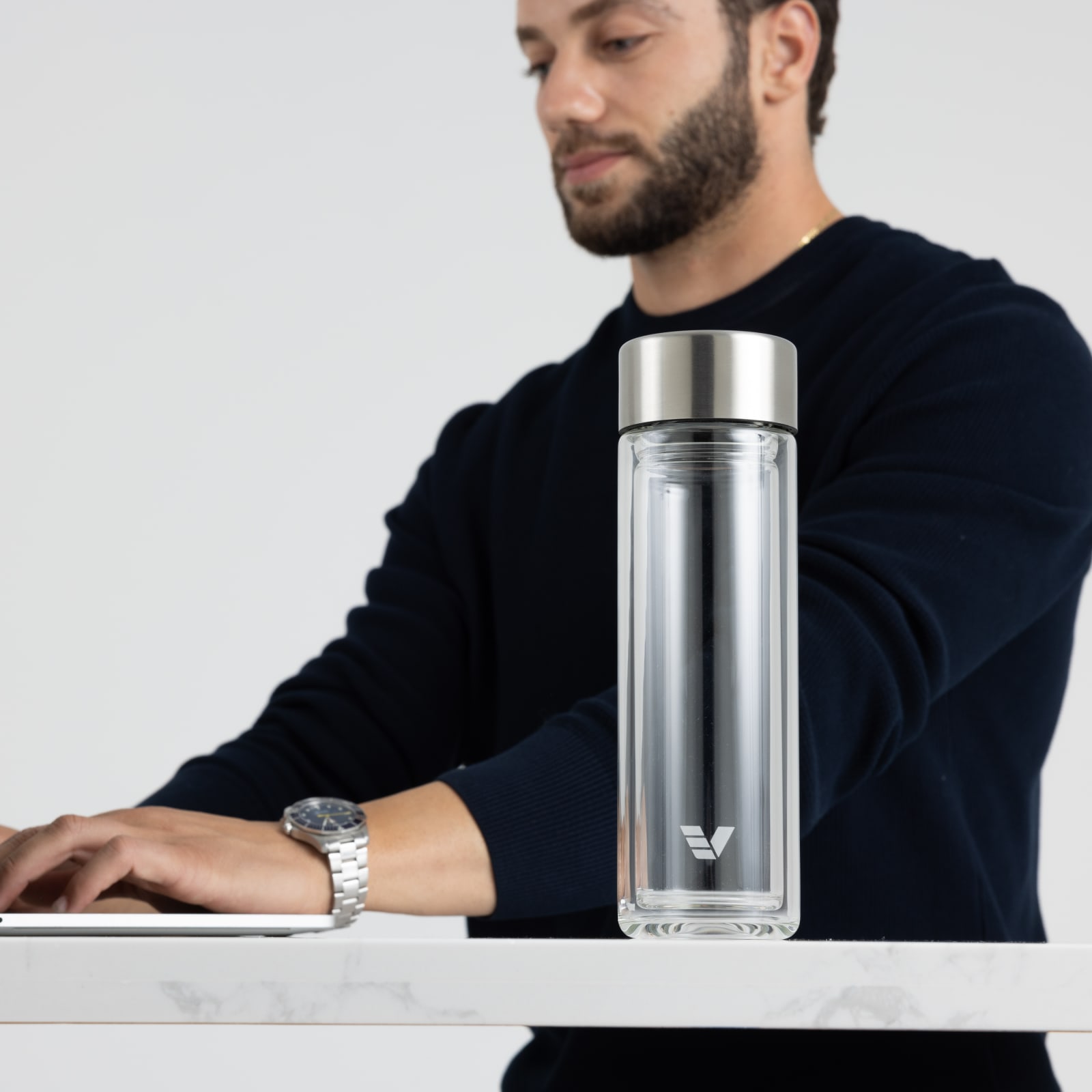 An Ever Vessel Glass Multi  double walled glass tea infuser water bottle with stainless grey lid sitting on a desk next to a male working.