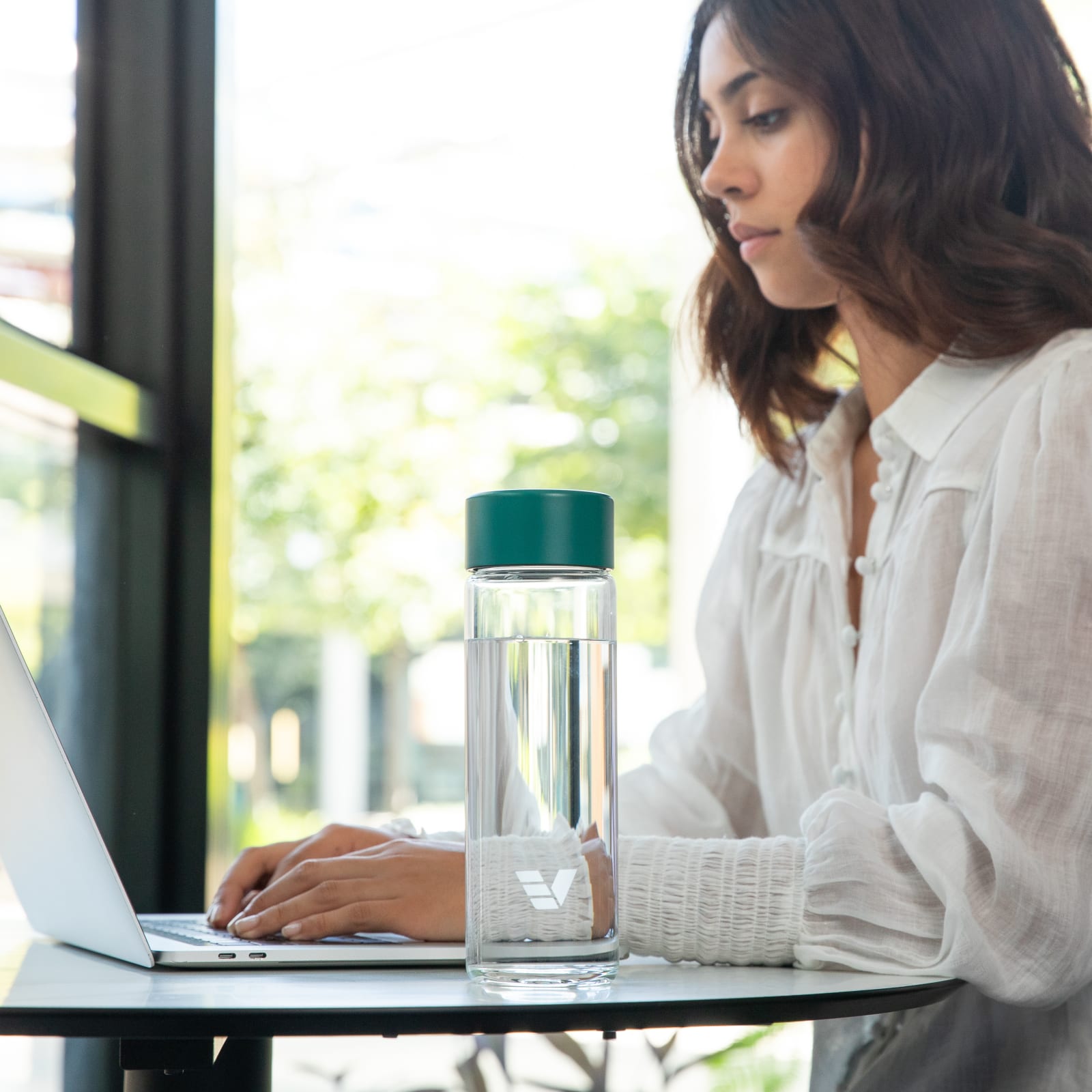 A female at a shared office desk with her Ever Vessel Glass water bottle with teal green lid by her side.