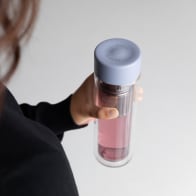Female holding a Personalized Ever Vessel Glass Multi double walled glass tea flask bottle with lilac colored lid and berry infused tea inside and matra engraved on the top.