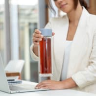 Female lifting an Ever Vessel Glass Multi double wall glass tea flask bottle with lilac colored lid and black infused tea inside at her desk.