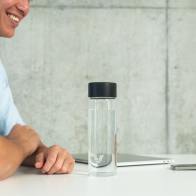 An Ever Vessel Glass water bottle with black lid on an open plan work desk next to a person talking to a work colleague.