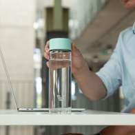 A male picking up his Ever Vessel Glass Multi tea infuser water bottle with a mint green lid off his desk.