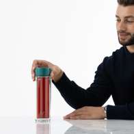 A male picking up his Ever Vessel Glass Multi  double walled glass tea infuser water bottle with a teal green lid and tea inside off his desk.