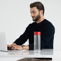 An Ever Vessel Glass Multi double wall glass tea infuser water bottle with red lid on stone desk next to a male working on his laptop.