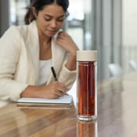 An Ever Vessel Glass Multi double wall glass tea infuser water bottle with beige lid and tea inside on a desk.