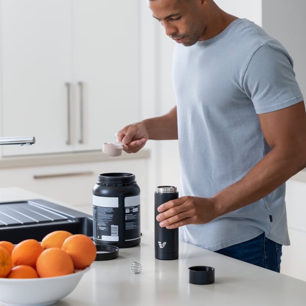 A male adding protein powder to his Ever Vessel Multi Black stainless steel tea infuser water bottle with whisk ball accessories adjacent on the kitchen bench.