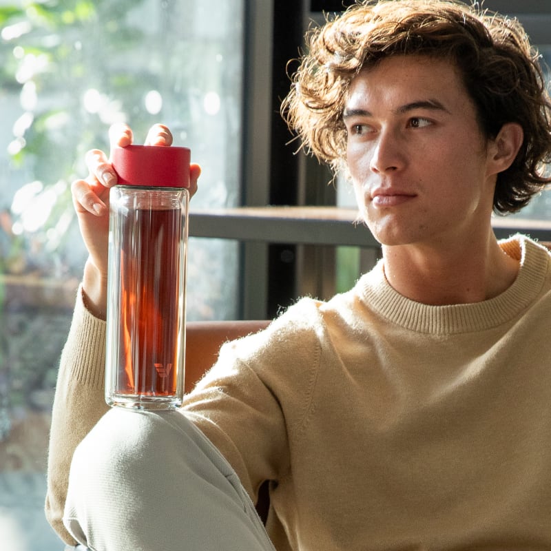 A male holding an Ever Vessel Glass Multi tea flask bottle with red colored lid and black infused tea inside on his knee.