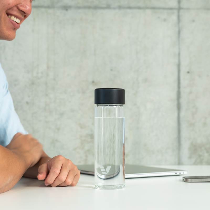 An Ever Vessel Glass water bottle with black lid on an open plan work desk next to a person talking to a work colleague.