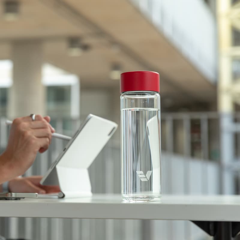 An Ever Vessel Glass water bottle with red lid on an open plan work desk next to a person working at his laptop.