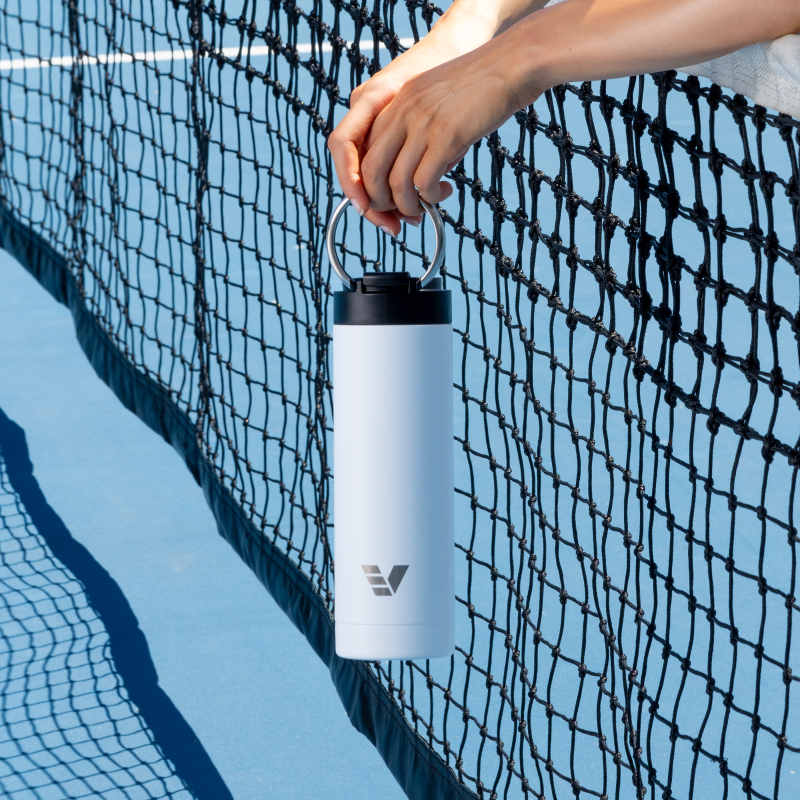 A female holding her Ever Vessel Maxi White Sand stainless steel water bottle by the integrated handle over the tennis court net.