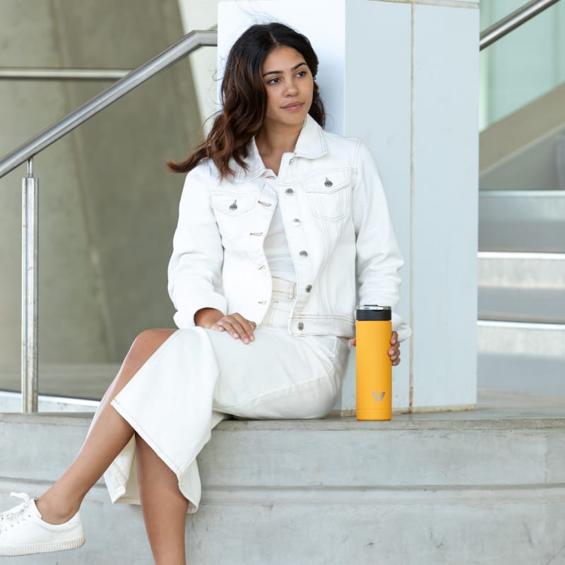 A female sitting next to her Ever Vessel Maxi yellow stainless steel water bottle outside of the work office.