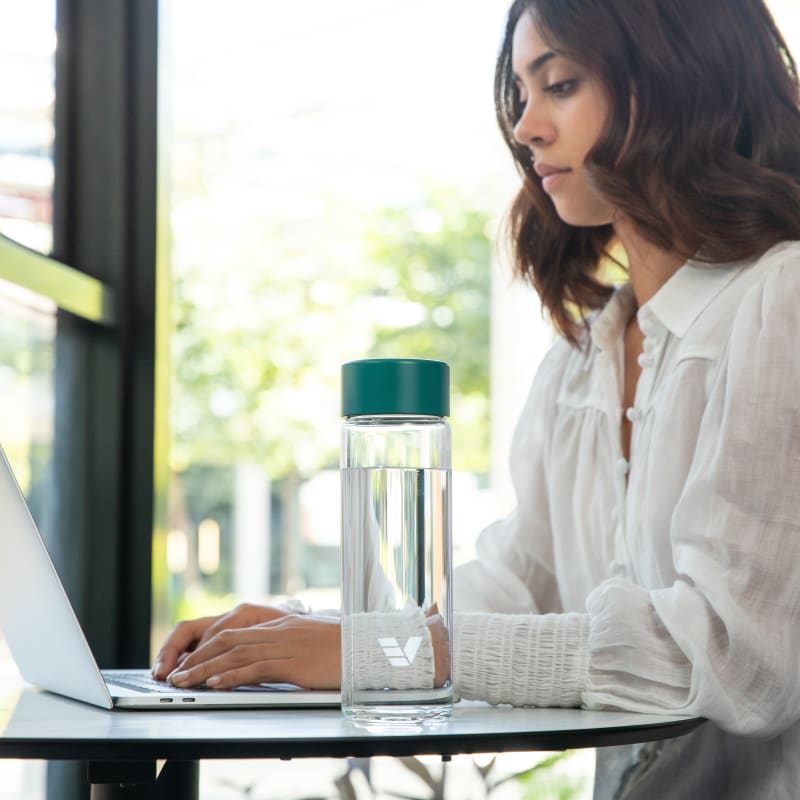 A female at a shared office desk with her Ever Vessel Glass water bottle with teal green lid by her side.