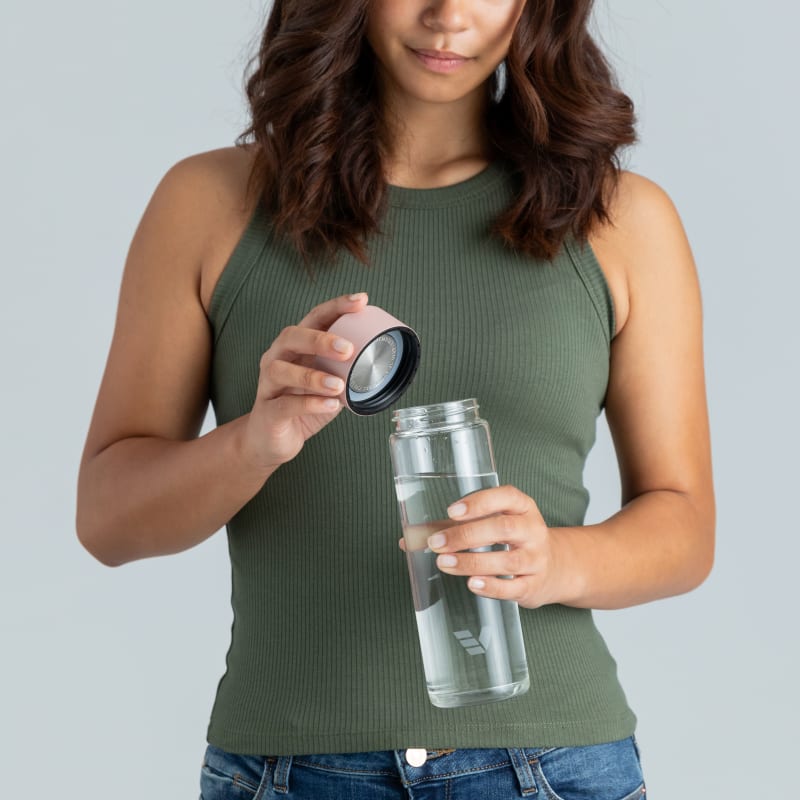 Female holding her optically clear borosilicate glass water bottle, with rose pink lid off at home.