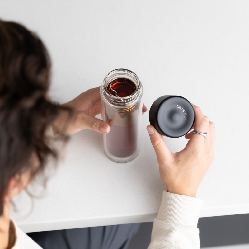 A male holding his Personalized Ever Vessel Glass Multi double walled glass tea infuser bottle with the black lid removed showing the engraved text 'Dad's Tea'.