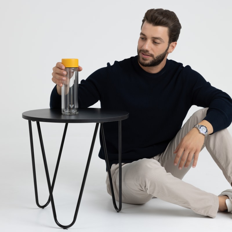 An Ever Vessel Glass Multi  double walled glass tea infuser water bottle with yellow lid sitting on a small table next to a seated male.