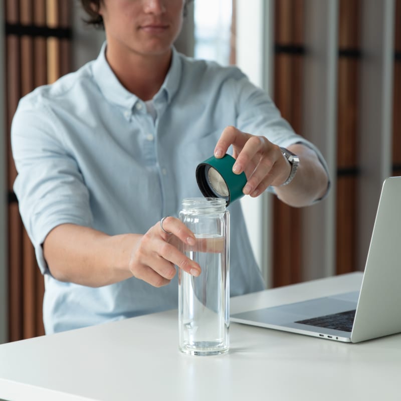 A male taking off the teal green lid from his Ever Vessel Glass Multi double walled glass tea infuser water bottle at his desk.